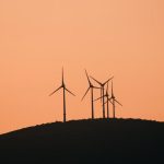 Wind turbines silhouetted against a vibrant yellow sunset sky on a rural hilltop.